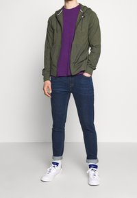 Zelená mikina na zip s předními kapsami, nošená přes fialové tričko. Kombinováno s modrými džínami a bílými teniskami s modrými detaily.