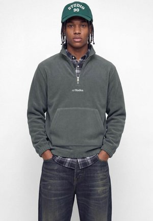 Jeune homme aux cheveux tressés portant une casquette "Studio 90" vert foncé, un sweat polaire gris JJ Studios à zip court, une chemise à carreaux et un jean foncé, les mains dans les poches.