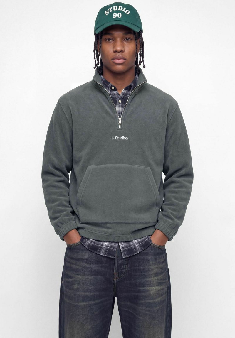 Jeune homme aux cheveux tressés portant une casquette "Studio 90" vert foncé, un sweat polaire gris JJ Studios à zip court, une chemise à carreaux et un jean foncé, les mains dans les poches.