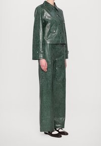 Zielona kurtka z eko skóry o skróconym kroju z ostrym kołnierzem i kieszenią na zamek, zestawiona z pasującymi szerokimi spodniami o błyszczącej fakturze.