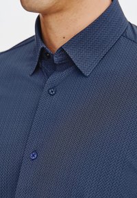 Granatowa koszula w wzory z kołnierzykiem zapinanym na guziki, teksturowana tkanina z subtelnym wzorem jodełki oraz niebieskie guziki.