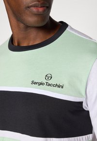 Zelené, čierne a biele pruhované tričko s okrúhlym čiernym golierom. Na zelenej časti je čierne logo "Sergio Tacchini".