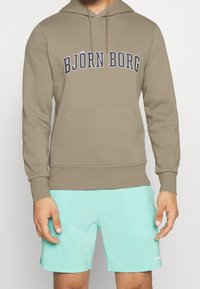 Hnědá mikina s kapucí se předními klokankovými kapsami a výrazným textem "BJÖRN BORG" na hrudi, zkombinována se světle modrými šortkami.