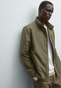 Giacca leggera verde oliva con colletto rialzato, bottoni a pressione e tasche laterali, indossata sopra un maglione grigio e pantaloni beige.