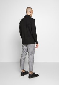 Man with tattoos wearing a black long-sleeve shirt, gray plaid pants, and black shoes stands facing a white wall.