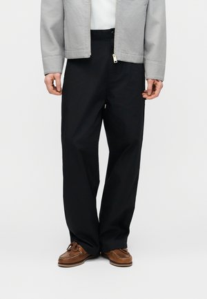 Férfi fekete bő szárú nadrágban, barna fűzős cipőben, világosszürke zakóban és fehér ingben, egyszerű világos háttér előtt.