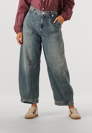 Weite blaue Jeans aus Denim mit einem verwitterten Look, ausgestattet mit zwei vorderen Taschen, einer hohen Taille und einem Knopfverschluss. Kombiniert mit cremefarbenen Sneakers.