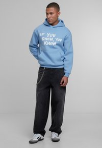 Svetlo modra kapuca z belim napisom "ČE VEŠ, VEŠ," v kombinaciji z ohlapnimi črnimi kavbojkami s ketno. Obuta z belimi supergami.