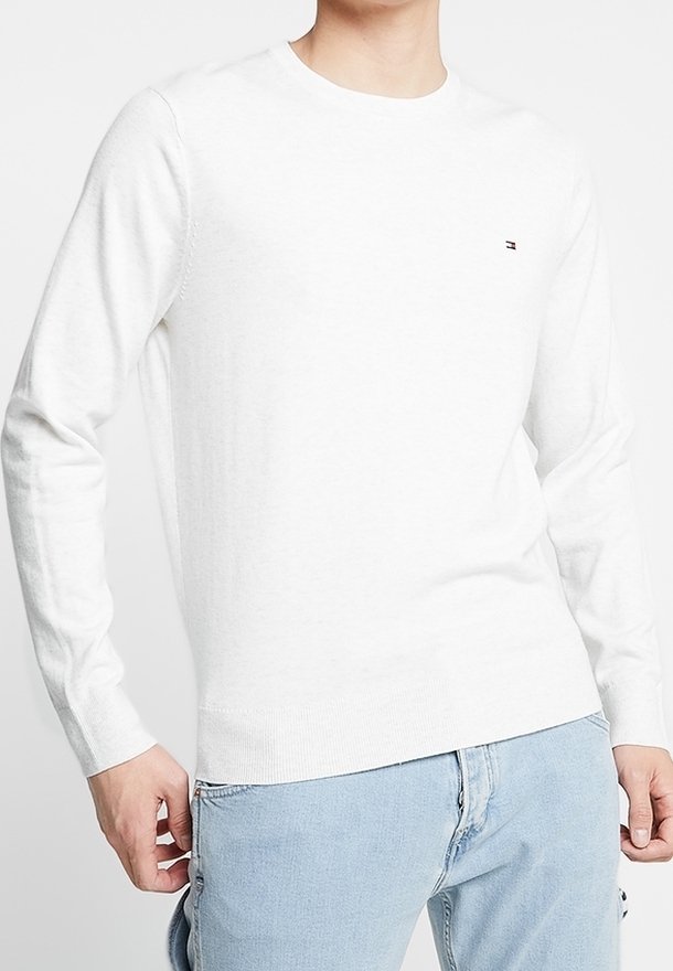 Homme portant un pull blanc uni à manches longues et un jean bleu clair, debout les mains près des poches sur un fond neutre.