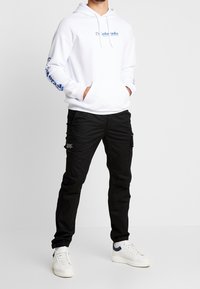 Witte hoodie met blauw logo op de voorkant en mouwen, gecombineerd met zwarte cargo pants met zijzakken en witte sneakers met blauwe accenten.