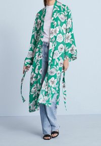 Blomstret grøn kimono med brede ærmer, prydet med hvide blomster og blade. Samlet med lyseblå jeans og sorte slidesandaler.