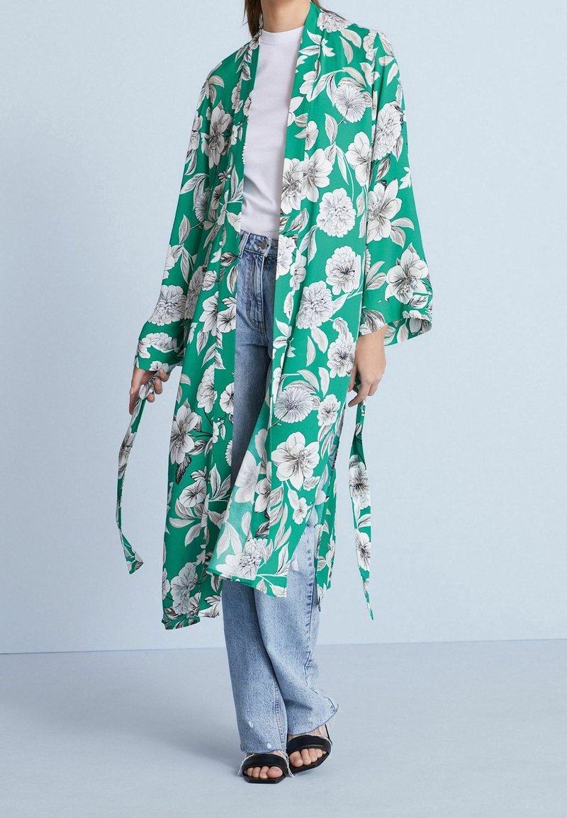 Blomstret grøn kimono med brede ærmer, prydet med hvide blomster og blade. Samlet med lyseblå jeans og sorte slidesandaler.