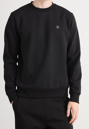 Osoba nosící černou mikinu s dlouhými rukávy a malým kruhovým logem na levé straně hrudníku, ruce uvolněné podél těla.