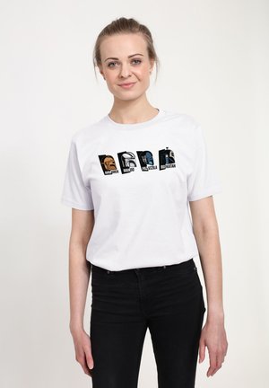 Kvinde med lyst hår iført en hvid t-shirt med stiliserede hjelme mærket Armorer, Mando, Paz Vizsla og Bo-Katan.