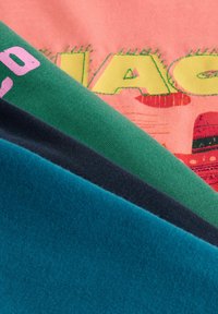 Gros plan sur des échantillons de tissu pliés en sarcelle, gris foncé et vert, posés sur un tissu rose avec des lettres brodées en jaune et rouge.