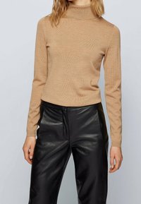 Suéter beige de cuello alto en material de punto suave con mangas largas, combinado con pantalones de cuero negros, que presentan un acabado liso y un ajuste relajado.