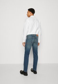 Dark blue denim jeans with a tapered fit, featuring two back pockets and a brown leather waistband accent, paired with a white shirt and black shoes.