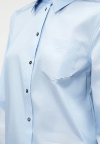 Camisa azul-clara de botões, feita de um tecido suave, com botões pretos, um bolso no peito costurado e um bordado de logotipo discreto.
