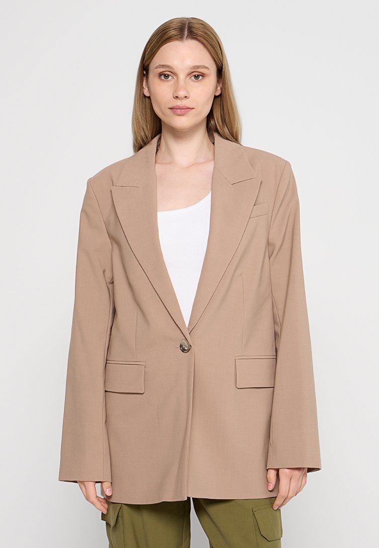 Giovane donna con capelli lisci castano chiaro che indossa un blazer beige oversize sopra una maglietta bianca e pantaloni cargo verde oliva, in piedi contro uno sfondo semplice.