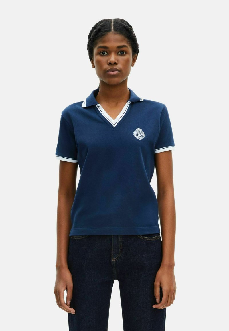 Marine polo skjorte med hvide accenter, korte ærmer og V-udskæring. Den har et broderet emblem på venstre bryst. Fremstillet af bomuld.