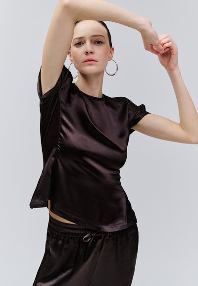 Femme aux cheveux tirés en arrière portant de grandes boucles d'oreilles en anneau et un haut à manches courtes en satin noir avec un pantalon assorti, levant les bras au-dessus de la tête.