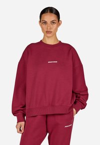 Žena oblečená v odpovídající burgundy oversized mikině a teplácích s bílými logy "PEGADOR", stojící proti jednobarevnému světlému pozadí.
