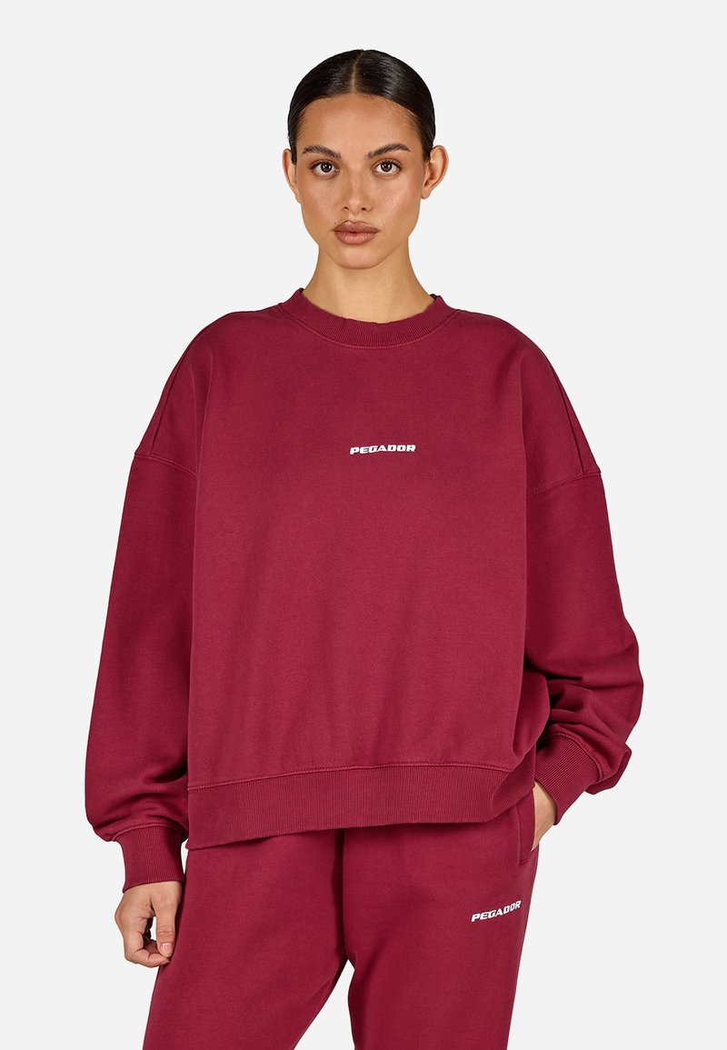 Žena oblečená v odpovídající burgundy oversized mikině a teplácích s bílými logy "PEGADOR", stojící proti jednobarevnému světlému pozadí.