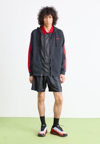 Hoodie preta com detalhes em vermelho, sobre uma camisa preta de padrão de manga curta e calções pretos. Usado com meias pretas e sapatilhas.