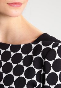 Kangas gaun hitam putih yang menampilkan pola polka dot besar. Lehernya lebar dengan tekstur yang halus.