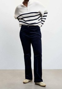 Weißer und marineblauer gestreifter Pullover mit einem halben Reißverschluss am Kragen, kombiniert mit marineblauen Cordhosen. Das Modell trägt beigefarbene Sneaker und steht vor einem neutralen Hintergrund.