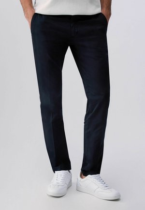 Uomo che indossa pantaloni neri aderenti e scarpe da ginnastica bianche, in piedi con le mani in tasca davanti a uno sfondo neutro.