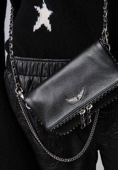 Borsa a tracolla in pelle nera con emblema d'argento a forma di ala e tracolla a catena, indossata sopra un maglione nero decorato con stelle e pantaloni dalla trama particolare.