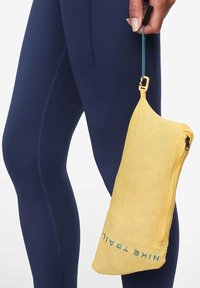 Funda amarilla con cremallera hecha de material texturizado, con una correa para sostener y el texto "Nike Trail" impreso en azul. Sostenida al lado de unos leggings azul marino.