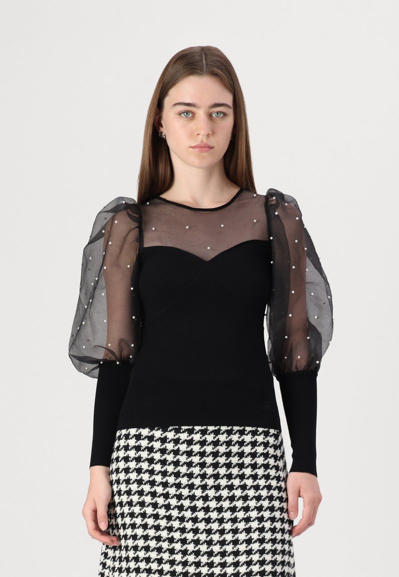 Zwarte top met doorzichtige, gepofte, gestructureerde mouwen versierd met witte kralen; aansluitende lijfje met een sweetheart-halslijn, gecombineerd met een houndstooth rok.
