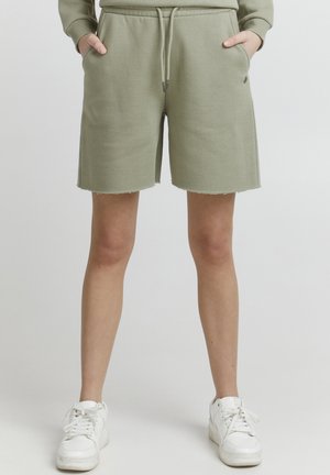 Pantaloni corti verde oliva, realizzati in tessuto morbido, con vita regolabile tramite un laccio, tasche laterali e orlo grezzo. Abbinati a sneaker bianche.