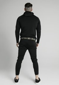 Sudadera atlética negra con joggers a juego. Presenta una capucha, diseño ajustado y una cinturilla con patrón. Textura suave, detalles mínimos.