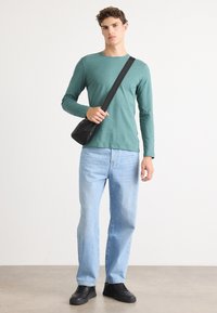Camicia verde a maniche lunghe, jeans blu chiaro a gamba dritta, borsa a tracolla nera e sneakers nere. Texture liscia, design casual.