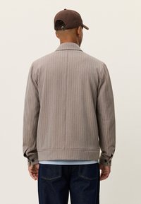 Καφέ σακάκι με ρίγες, γιακά, μακριά μανίκια και μανσέτες με κουμπιά. Φορεμένο πάνω από πουκάμισο ανοιχτού μπλε χρώματος, σε συνδυασμό με σκούρο τζιν.