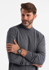 Šedý rolák z měkké tkaniny. Model má na sobě metalické hodinky, paže zkřížené, na čistém bílém pozadí.