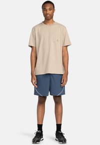 Beige Baumwoll-T-Shirt mit einer Brusttasche, kombiniert mit marineblauen Shorts mit geschwungenem weißen Piping. Tragen mit schwarzen Sportschuhen.