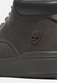 Bota de cuero gris con un acento de cuero negro, que presenta costuras grises y un logo de árbol embosado en forma de círculo en el lateral.