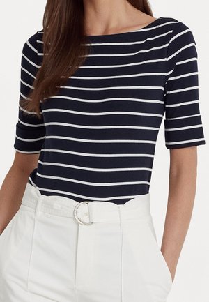 Femme portant un haut à rayures bleu marine et blanc à encolure bateau avec des manches aux coudes, rentrée dans un pantalon blanc taille haute avec une ceinture.