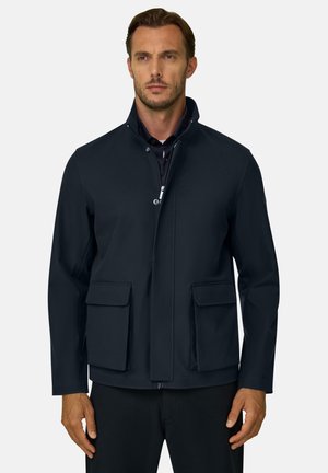 Mann trägt eine schwarze Jacke mit Reißverschluss, hohem Kragen und zwei großen vorderen Klappentaschen, steht vor einem schlichten Hintergrund.