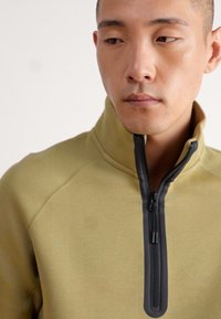 Αυτό το ελαιόχρωμο φούτερ με τσέπη σε σχήμα τετάρτου διαθέτει μαύρο φερμουάρ και ανυψωμένο γιακά, κατασκευασμένο από μαλακό, υφασμάτινο υλικό με εμφανείς ραφές.