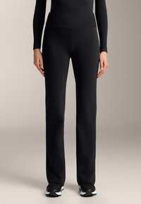 Čierne športové nohavice s širokým pásom, vyrobené z hladkej, elastickej tkaniny. Spárované s čiernymi teniskami s bielymi akcentmi.