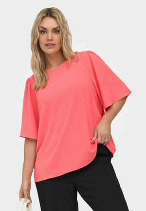 Blonde Frau trägt locker sitzendes korallenrosa Kurzarm-Top und schwarze Hose, hält ein weißes Tuch in der linken Hand und steht vor hellem Hintergrund.