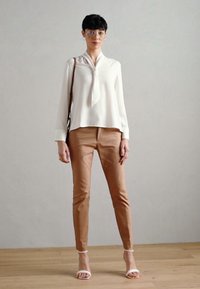 Weißes Bluse mit Bindekragen, beigefarbene maßgeschneiderte Hose und minimalistische weiße Sandalen. Besitzt zarte Manschetten und einen lockeren Schnitt. Steht auf Holzboden.