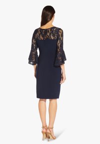 Tmavě modré šaty s krajkovými zvonovými rukávy, přiléhavým střihem a délkou po kolena. Krajka má květinový vzor, měkkou texturu a otevřený detail na zádech.