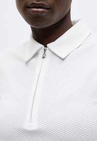 Άτομο με σκούρο δέρμα που φοράει λευκή πόλο μπλούζα με υφή και ασημένιο φερμουάρ που είναι μερικώς κλειστό κοντά στον γιακά.