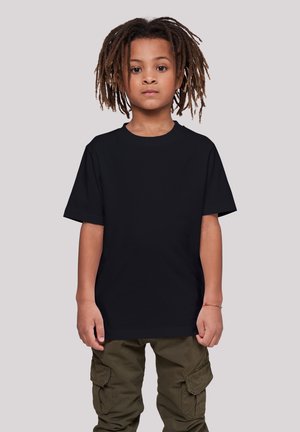 Child with medium brown skin and dreadlocks stands facing forward wearing a plain black t-shirt and olive green cargo pants against light background.
